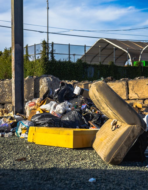 An outdoor scene showing a pile of mixed household waste and rubbish on the ground next to a low stone wall. The waste includes black and clear plastic bags filled with trash, a yellow plastic container, and a large, old, beige car seat with fabric upholstery that appears dirty and worn. The area is on a gravel or asphalt surface, with a metal pole and power lines visible in the background. Behind the wall, there are trees, a hedge, and a partially visible structure with a curved roof, suggesting a semi-urban environment. The scene is illuminated by natural daylight, highlighting the disorderly accumulation of waste, which may indicate an off-site private disposal or rubbish collection process managed by waste removal services like Waste Collection Paddington. This type of visible waste accumulation typically prompts the need for professional rubbish removal to ensure proper clearance and environmental responsibility.
