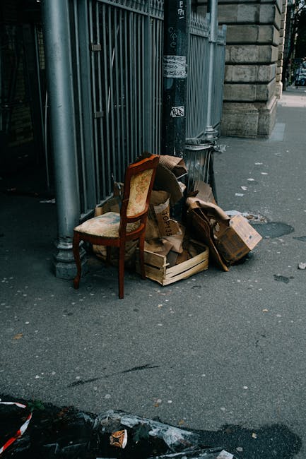 A daytime street scene showing a small pile of discarded waste items placed on the sidewalk next to a stone building and a metal fence. The waste includes a vintage-style upholstered wooden chair with a patterned fabric seat and backrest, which is positioned upright, and several brown cardboard boxes, some crushed or partially collapsed, along with a few loose pieces of packaging. The waste appears to be gathered for private collection or rubbish removal, situated on a paved footpath beside a stone wall and metal gate, with a black trash bag visible behind the cardboard boxes. The surrounding environment suggests an urban area, with a partial view of a street and pedestrians in the background. The scene conveys typical private or independent rubbish disposal, potentially managed by services like Waste Collection Paddington, and highlights the scope of non-public waste handling options involving on-site clearing of bulk furniture and packaging materials. The lighting is neutral, with natural daylight illuminating the scene, emphasizing the textures of the materials and the setting.