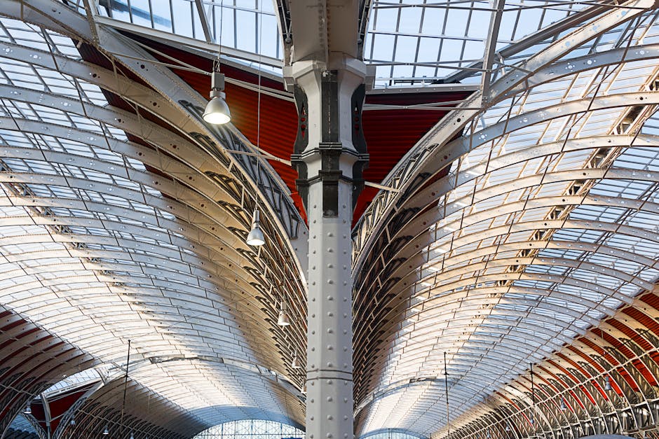 The image depicts the interior roof structure of a large commercial building, characterized by an extensive series of curved, metallic arches and beams that form a symmetrical, tunnel-like ceiling. The superstructure is composed of shiny metal panels with a reflective surface, showcasing a combination of silver and grey tones, and arranged in a layered pattern that emphasizes the architectural design. The ceiling integrates transparent glass sections allowing natural daylight to illuminate the space, complemented by industrial-style pendant lamps hanging from the beams, providing additional lighting. A central supporting column made of riveted steel runs vertically through the middle of the scene, dividing the view into two mirrored halves. The environment appears to be part of a spacious, indoor setting, possibly a warehouse or train station, with a focus on the structural elements and the interplay of light from above. This architectural detail can be associated with the careful design of large spaces that may require efficient indoor waste management or periodic on-site clearance, relevant to services like waste collection and rubbish removal by companies such as Waste Collection Paddington.