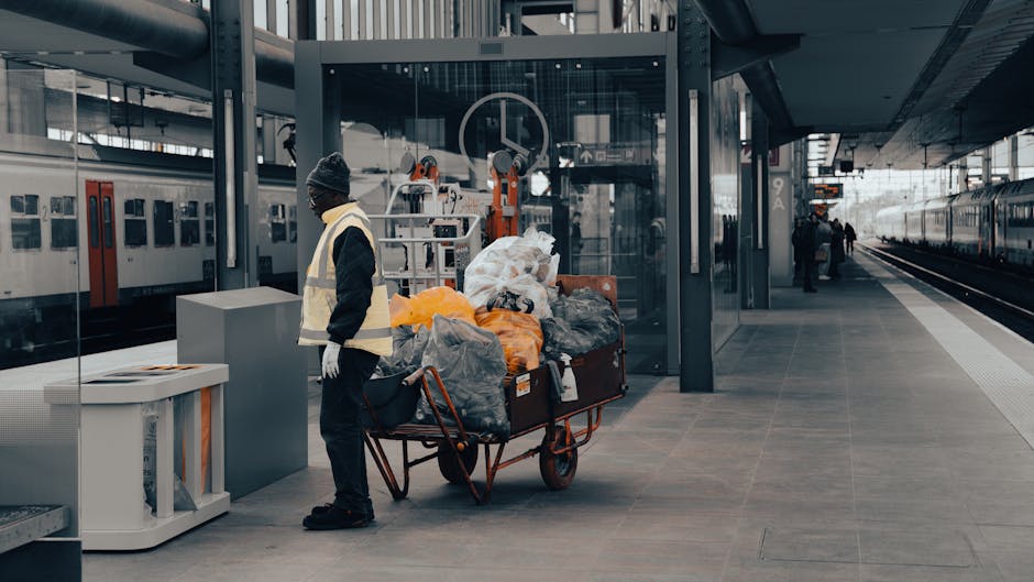 A man wearing a dark jacket, grey beanie, and white gloves stands next to a large red wheeled trolley loaded with various waste sacks and materials, including black, grey, orange, and white bags, some of which are partially open revealing loose rubbish. The trolley is positioned on a paved platform in an indoor train station with modern architectural elements, including glass walls and metal structural supports. In the background, there are trains on adjacent tracks, with a few passengers walking further along the platform. Nearby, a white waste bin with orange reflective bands is placed to the left of the man, and a tall grey column is situated behind him. The overall scene appears to depict an instance of on-site waste handling or collection, consistent with the services provided by Waste Collection Paddington in handling commercial or domestic rubbish removal at transit hubs.