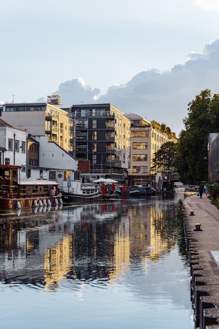 A daytime scene along a narrow canal featuring modern residential buildings with multiple floors, constructed from a combination of light-colored and dark materials, with large windows and balconies. In the foreground, a wooden boat docked alongside the canal shows a weathered texture, with some red accents on parts of the boat structure. To the right, a paved walkway runs parallel to the water, with a few bollards or mooring posts visible along the edge. The canal water reflects the buildings and partly cloudy sky above, creating a mirror-like surface. A large tree with dense green foliage is positioned on the right side, partially obscuring some of the buildings, while a person can be seen walking further along the path. The scene is illuminated by natural daylight, with the soft light highlighting the textures of the boat's wooden surface and the water. The overall setting suggests an urban waterside area, where private boating and waterfront living are common, and may relate to alternate waste collection options provided by Waste Collection Paddington, as indicated by the nearby boats and dock infrastructure.
