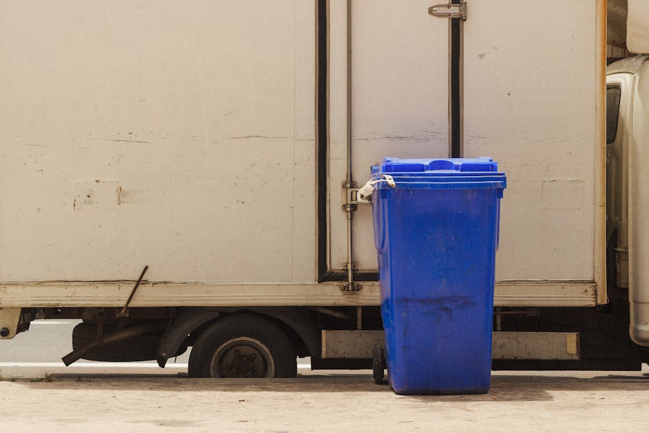 A tall, bright blue waste bin with a closed, ribbed lid is positioned on a concrete pavement next to a white delivery or service vehicle. The bin shows faint scuff marks and slight dirt on its surface. The vehicle, with a smooth, slightly weathered finish, features a large side panel, and is elevated on small wheels that are partially visible beneath the vehicle’s body. A vertical metal pipe extends from the top to the bottom of the vehicle’s side, secured with metal brackets. The background consists of a plain, pale wall with minor surface imperfections, and a closed black door with a metallic handle is visible behind the vehicle. The scene is evenly lit, suggesting natural daylight, and the setting appears to be a typical urban environment where private waste collection or on-site clearance services, like those provided by Waste Collection Paddington, might operate to manage rubbish collection outside residential or commercial properties.