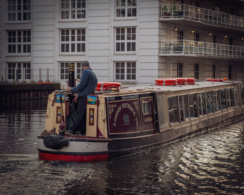 A daytime scene along a narrow canal featuring modern residential buildings with multiple floors, constructed from a combination of light-colored and dark materials, with large windows and balconies. In the foreground, a wooden boat docked alongside the canal shows a weathered texture, with some red accents on parts of the boat structure. To the right, a paved walkway runs parallel to the water, with a few bollards or mooring posts visible along the edge. The canal water reflects the buildings and partly cloudy sky above, creating a mirror-like surface. A large tree with dense green foliage is positioned on the right side, partially obscuring some of the buildings, while a person can be seen walking further along the path. The scene is illuminated by natural daylight, with the soft light highlighting the textures of the boat's wooden surface and the water. The overall setting suggests an urban waterside area, where private boating and waterfront living are common, and may relate to alternate waste collection options provided by Waste Collection Paddington, as indicated by the nearby boats and dock infrastructure.