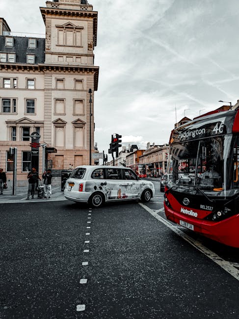 A busy urban street scene in Paddington during daytime, featuring a white chauffeur-driven car with a sleek exterior and dark tinted windows positioned at a pedestrian crossing, with a contemporary red and black double-decker bus marked with route information nearby. In the background, a historic beige stone building with detailed architectural features, including decorative cornices and window surrounds, is partly visible, alongside various storefronts and traffic signals. Several pedestrians are waiting to cross the road, some with shopping bags, while others are engaged in conversation. The sky above is overcast with light grey clouds, casting diffuse lighting over the scene. The road surface appears clean and well-maintained, with lane markings and curb edges clearly visible, indicating a central urban area that relies on both on-street parking and official public transport. This setting subtly relates to the broader context of city transportation and can be associated with alternative waste management options by highlighting the importance of efficient urban logistics, including private vehicle use and public transit, in supporting integrated waste collection services like those offered by Waste Collection Paddington.