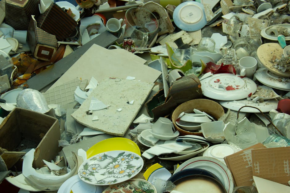 An extensive heap of discarded household items and broken crockery, comprising ceramic plates, bowls, mugs, and various kitchenware, is spread across a surface such as a driveway or outdoor area. The ceramics feature different colours and finishes, including plain white, floral patterns, and multicoloured designs, with many shards and chipped pieces visible. Among the debris, there are also miscellaneous materials like cardboard boxes, a piece of foam or insulation, and plastic items mixed in with the ceramic waste. The pile appears chaotic with items stacked, leaning, or partially buried under other rubbish, and some ceramics show signs of damage or breakage. The environment is outdoors, illuminated by natural daylight, highlighting the complexity and volume of the waste, which is typical of an on-site clearance or private waste disposal scenario undertaken by a professional waste management service such as Waste Collection Paddington. This scene illustrates the type of large-scale, mixed waste that might be collected during an independent rubbish removal or builder’s waste cleanup effort.