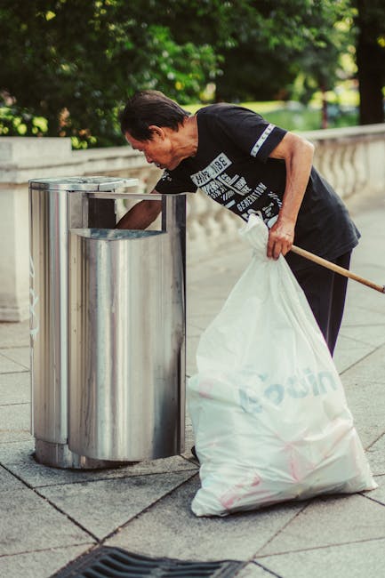 A middle-aged man with dark hair, dressed in a black T-shirt with white graphic text on the front, is seen standing on a paved pedestrian area beside a stone railing, next to a stainless steel public trash bin. He is holding a large white rubbish bag, which appears to be filled with waste, and is leaning forward, actively depositing waste into the bin. The scene is outdoors, with lush green trees and foliage in the background, suggesting a park or city street environment. The man is using a wooden stick or pole to assist with rubbish collection, indicating a form of street cleaning or a community cleanup effort. The stainless steel bin has a modern, cylindrical design with an opening at the top, located on a concrete surface that is clean and well-maintained. The overall setting conveys an emphasis on waste management and rubbish disposal, aligning with services like independent rubbish collection or street waste clearance, as provided by Waste Collection Paddington.