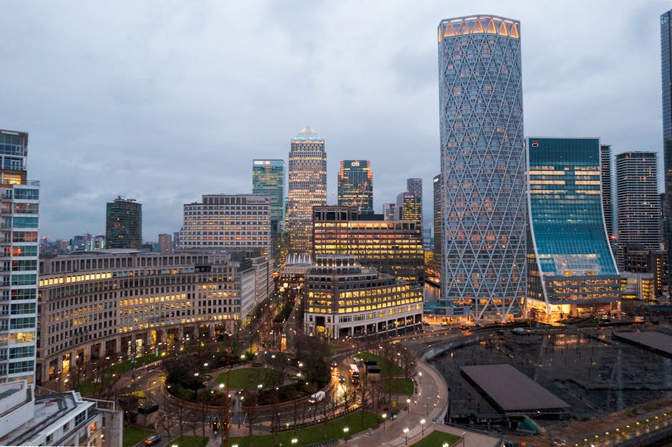A cityscape featuring a cluster of modern office buildings and skyscrapers, primarily constructed from glass, steel, and concrete, with reflective windows and sleek facades. The tallest building on the right has a distinctive crisscross patterned exterior with a curved outline, illuminated by warm ambient lighting. Several of the lower buildings display illuminated windows with yellow and white lights, indicating active offices or commercial spaces. In the foreground, a small urban park is visible with a walking path, manicured lawns, and leafless trees, suggesting a late autumn or winter setting. Surrounding the park, a street with moving vehicles has their headlights and taillights creating streaks of light on the wet pavement. The overall scene is set during dusk or early evening with a cloudy sky casting a muted, diffused light over the city. This urban setting exemplifies contemporary office and commercial development in a financial district, with Waste Collection Paddington providing private waste disposal services in areas like Sheldon Square and W2, as part of alternative waste handling options for local businesses.