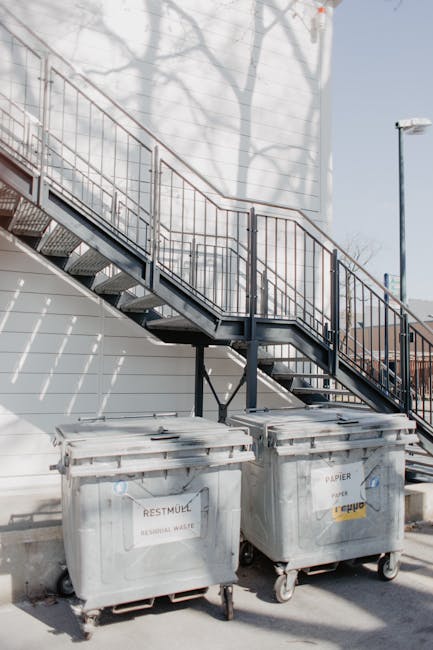The image depicts two large, grey wheeled waste bins positioned in front of a white building with horizontal siding. The bins are labeled for different waste types; the left one is marked for residual waste, while the right one is designated for paper, with visible text indicating its purpose for recyclable paper waste. The bins are made of plastic with a textured surface and are placed directly on a paved surface. Behind the bins, a black metal fire escape staircase with a grated platform and railings extends diagonally upward along the building's exterior wall. Shadows of leafless tree branches are cast onto the building's surface, suggesting a sunny day, and a streetlamp is visible in the background, to the right of the staircase. The setting appears to be an urban area, consistent with private waste collection or rubbish removal services such as those provided by Waste Collection Paddington, supporting alternative waste handling outside of council collection systems.