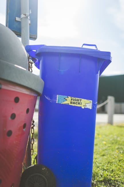 A tall, rectangular blue plastic rubbish bin with a closed lid, positioned outdoors on a paved area, slightly to the right of the image. The bin has a small, rectangular yellow and black sticker with the words 'FIGHT BACK!' affixed to its front. To the left, part of a red plastic bin with round ventilation holes and a black lid is partially visible, with a black wheel at the bottom for mobility. In the background, a parking lot or street scene is visible, with a blue sign mounted on a metal pole, some green grassy patches, and a white fence or barrier. The scene is illuminated by natural daylight, suggesting a bright, clear day, and the overall environment appears to be part of a waste collection area or roadside bulk refuse collection spot, typical for private rubbish removal services like Waste Collection Paddington, in a suburban or urban setting.