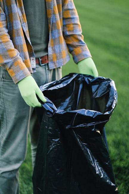 A person standing outdoors on a grassy area holding open a large black plastic rubbish bag, which appears to be partially filled with waste. The individual is dressed in a grey shirt layered with a yellow and blue checkered flannel shirt, and is wearing bright green gloves for handling waste materials. The background shows a well-maintained, green lawn, indicating a garden or park setting. The scene suggests an activity related to waste collection or outdoor rubbish clearance, with the individual preparing to dispose of general household or garden refuse. The image emphasizes the preparation involved in independent waste removal, highlighting the use of suitable protective gloves and the handling of large, durable rubbish bags, which is often part of private or alternative waste handling services carried out by companies such as Waste Collection Paddington. The natural lighting and clear environmental context support visual clarity and accessibility, providing a straightforward depiction of rubbish collection in an outdoor environment.