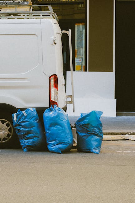 A dark red flatbed pickup truck parked on an urban street with a modern office building in the background. The truck's cargo area is filled with various types of rubbish, including black and white plastic bags, some of which are partially open, revealing debris inside. The truck features a protective metal grid at the cab's rear window and small rear lights, with the tailgate lowered to accommodate the loaded waste. The vehicle appears to be involved in private rubbish collection or on-site clearance services provided by Waste Collection Paddington, suitable for alternative waste handling outside of local authority disposal. The scene is outdoors in daylight with natural, even lighting, and the environment shows a city environment with the building's large windows and smooth concrete surfaces behind the vehicle. The image captures an example of independent rubbish removal activity typically associated with professional waste management services in busy urban areas, emphasizing the effective collection of waste for disposal or recycling.