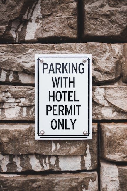 A rectangular metal sign with a white background and black text that reads 'PARKING WITH HOTEL PERMIT ONLY' is fixed to a textured stone wall composed of large, irregularly shaped stones with a rough surface and natural earthy tones, including shades of brown, beige, and gray. The sign is secured at each corner with visible round-headed screws, and it provides clear instructions limiting parking to vehicles displaying a valid hotel permit. The surrounding environment suggests an exterior setting, likely on a building facade or boundary wall, with no other objects or fixtures visible in the immediate vicinity. The lighting appears natural, contributing to the clarity of the sign and the details of the stone wall behind it, supporting the context of designated parking restrictions in a private or commercial property area managed by an organisation such as Waste Collection Paddington for waste management or property regulation enforcement related to on-site or private waste disposal procedures.