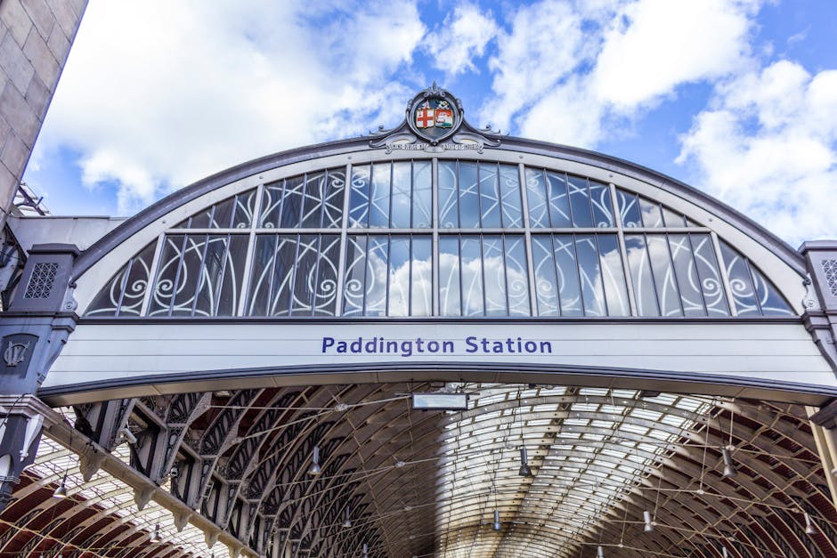 A close-up view of the entrance archway of Paddington Station, featuring an ornate, semi-circular metal framework with decorative scrollwork and lattice patterns painted in dark grey or black. The arch is topped with a crest emblem displaying a shield with a red cross and a blue background, positioned centrally at the apex. Below the arch, a white signage panel displays the name 'Paddington Station' in blue lettering. The background shows a partly cloudy sky with patches of blue and white clouds, illuminating the station's intricate ironwork and glass roofing structure. The lower section of the image reveals the station's covered platform area with metal support beams and a curved glass canopy that allows natural light to filter through, creating a well-lit environment suitable for passengers and staff. The overall scene emphasizes the architectural detail and classical design elements characteristic of historic railway station entrances, highlighting the station's exterior as a notable urban landmark associated with transportation and travel in London.