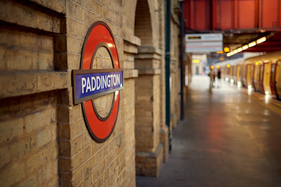 A close-up view of a circular London Underground station sign mounted on a textured, yellowish-brown brick wall at Paddington station. The sign features a red outer ring, a white inner circle, and a blue rectangular panel in the center with the word 'PADDINGTON' in bold white letters. To the right of the sign, the station platform extends into the background, with a row of orange and yellow tiled walls and a set of train doors partially visible. Soft, warm lighting illuminates the platform, creating a contrast with the darker, dimly-lit background where a few people can be seen walking or standing. The environment suggests a busy urban transportation hub, with the station sign serving as a key visual cue for travelers. Waste Collection Paddington’s rubbish removal services could be relevant in managing waste generated by station operations, although this is not directly depicted in the image. The scene captures the typical atmosphere of an underground railway station, with detailed textures of brickwork, station signage, and tiled surfaces, emphasizing the functional environment of public transportation and associated waste management considerations.